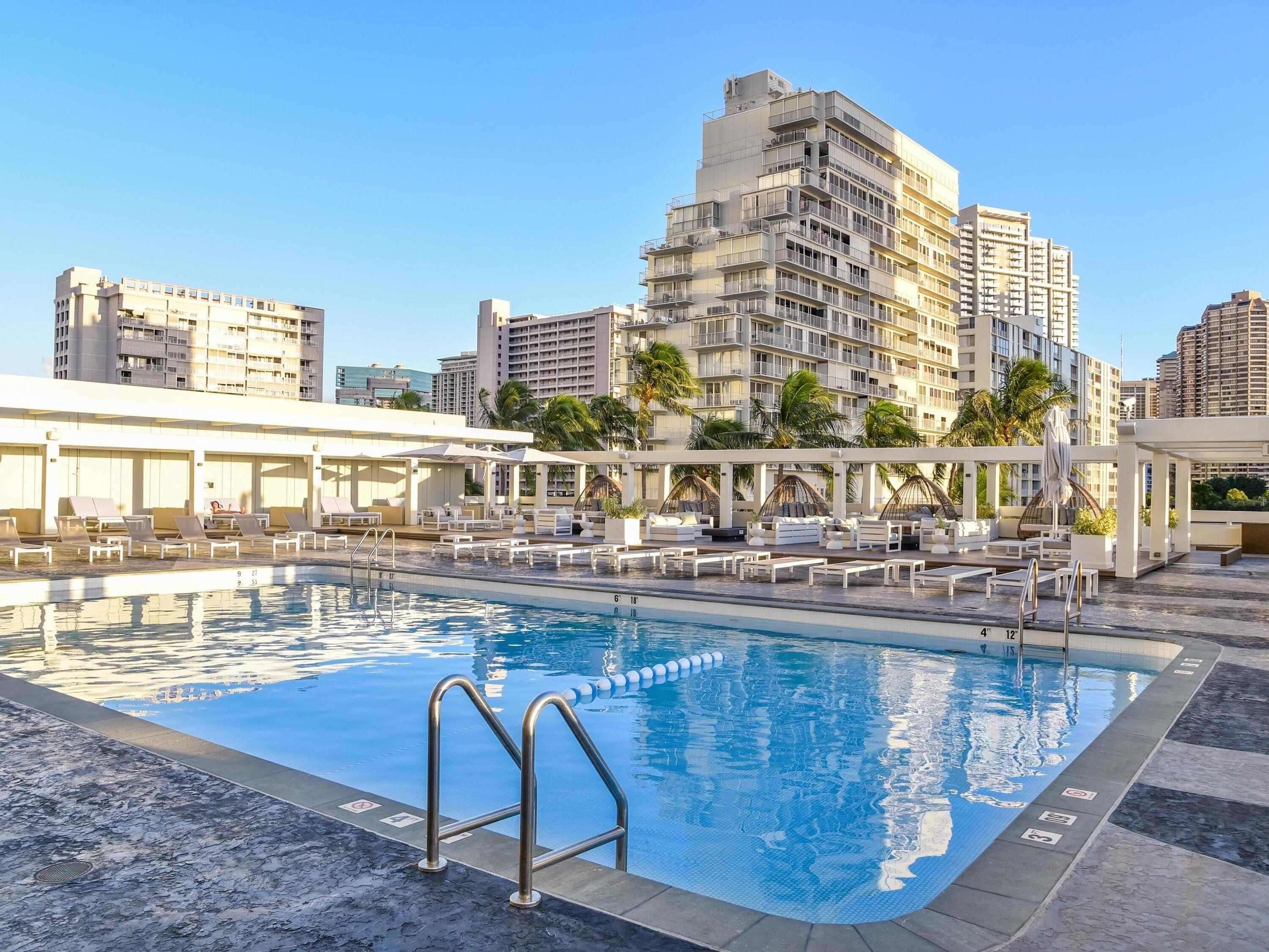 A rooftop pool with lounge chairs and umbrellas, surrounded by high-rise buildings.