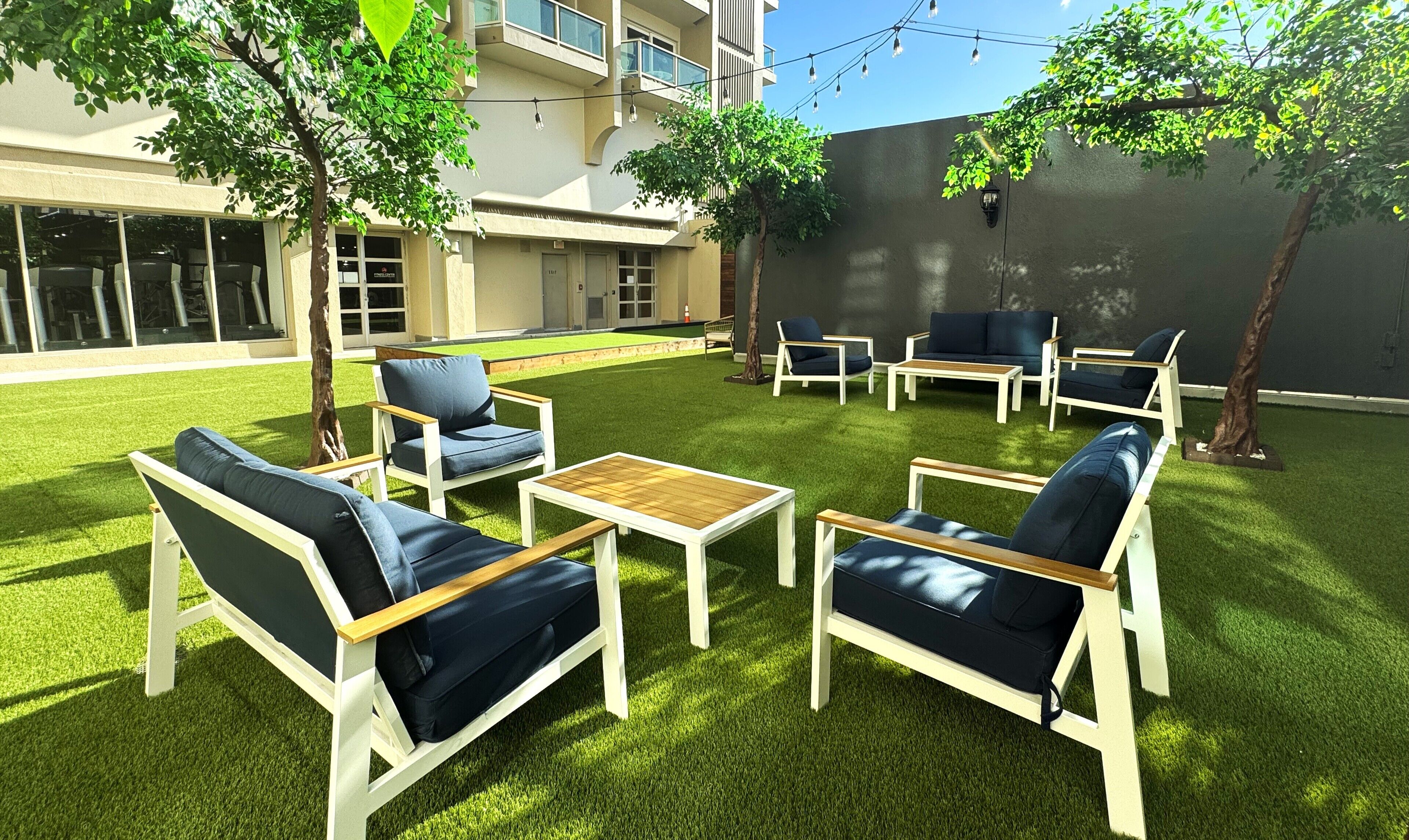 An outdoor seating area with white and blue furniture, surrounded by artificial grass and trees.