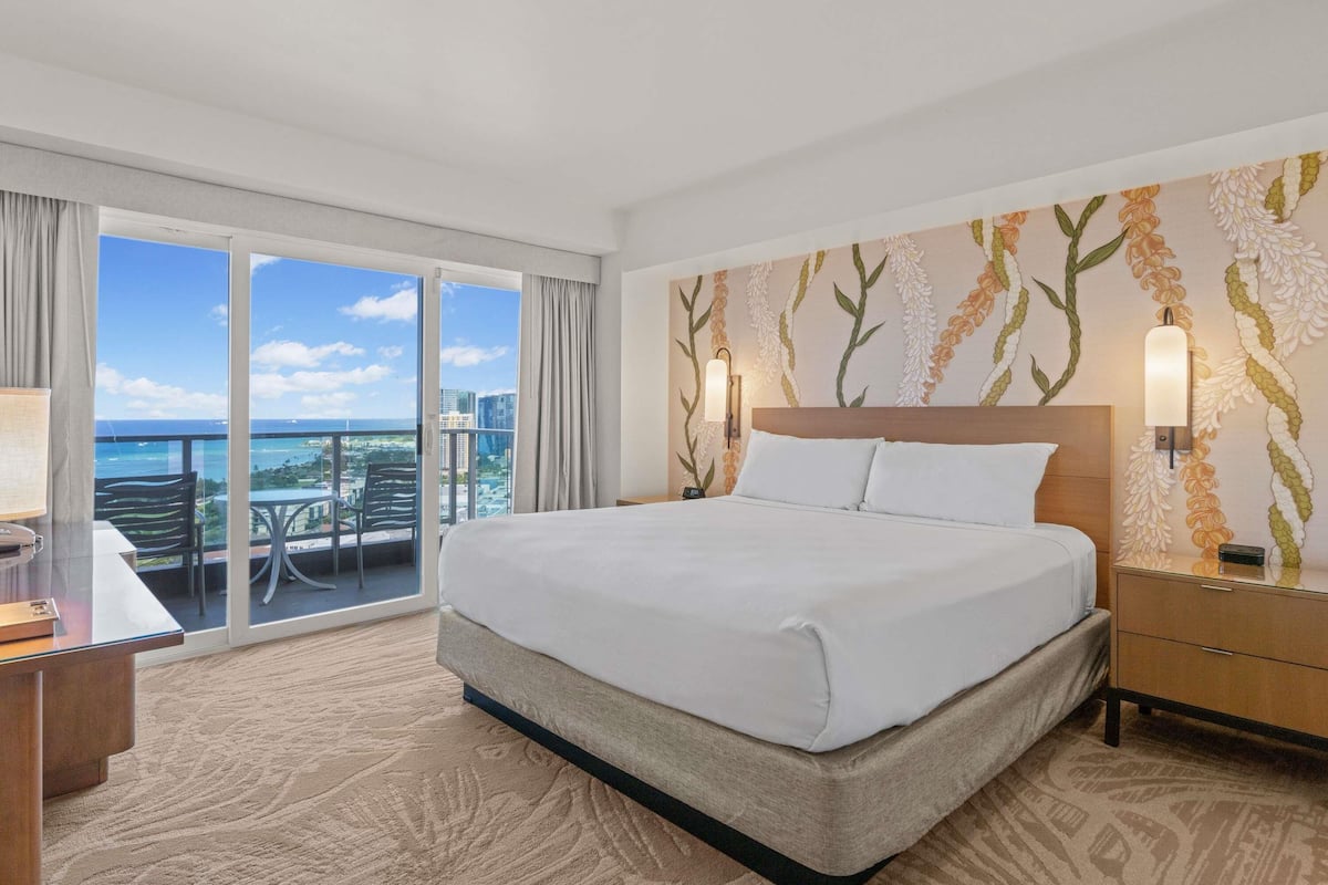 Habitación de hotel con cama, escritorio, silla, mesita de noche y vista al océano.