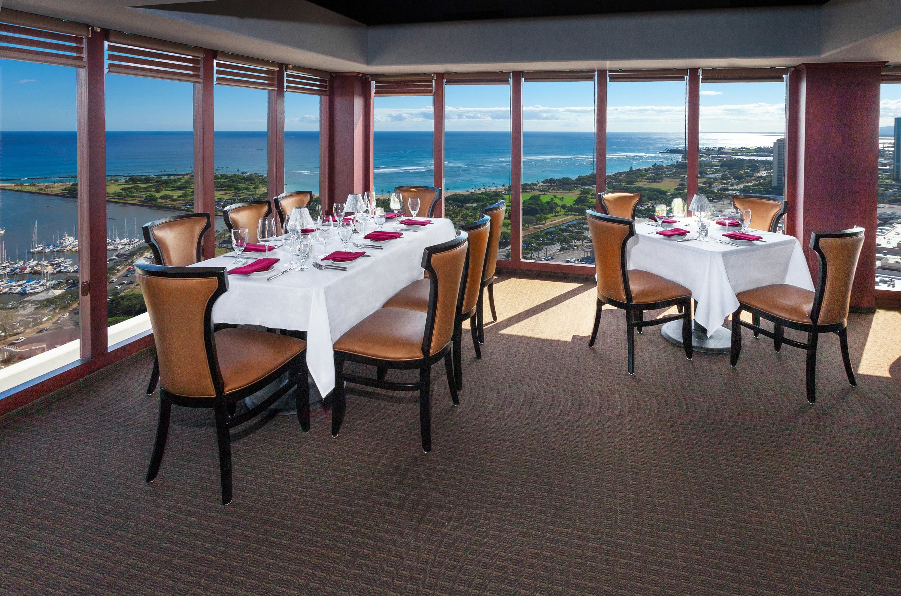 A dining area with tables set for four, large windows offering a view of the ocean and marina.