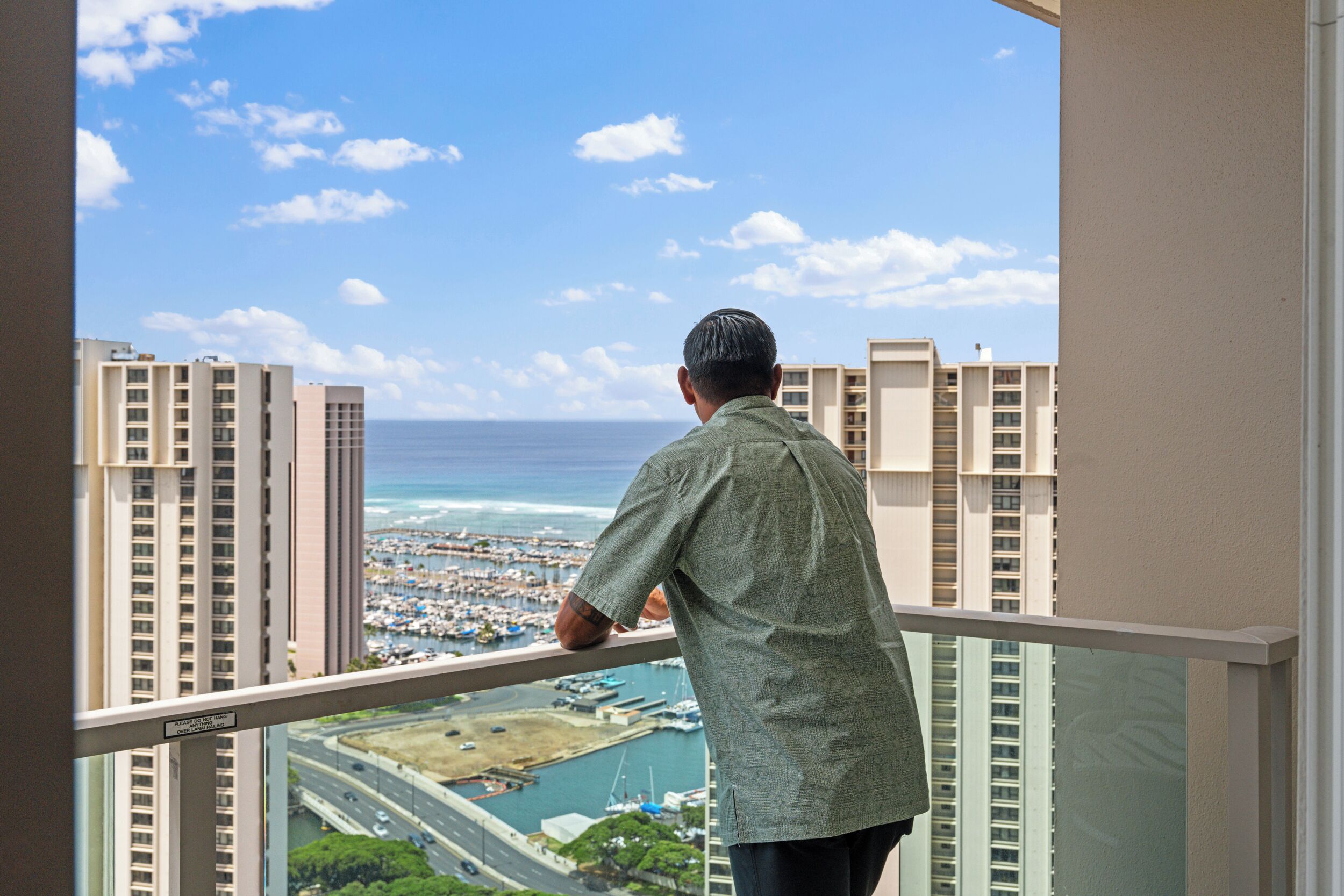 Um homem em uma varanda, com vista para um horizonte costeiro com edifícios altos e um porto de iates.