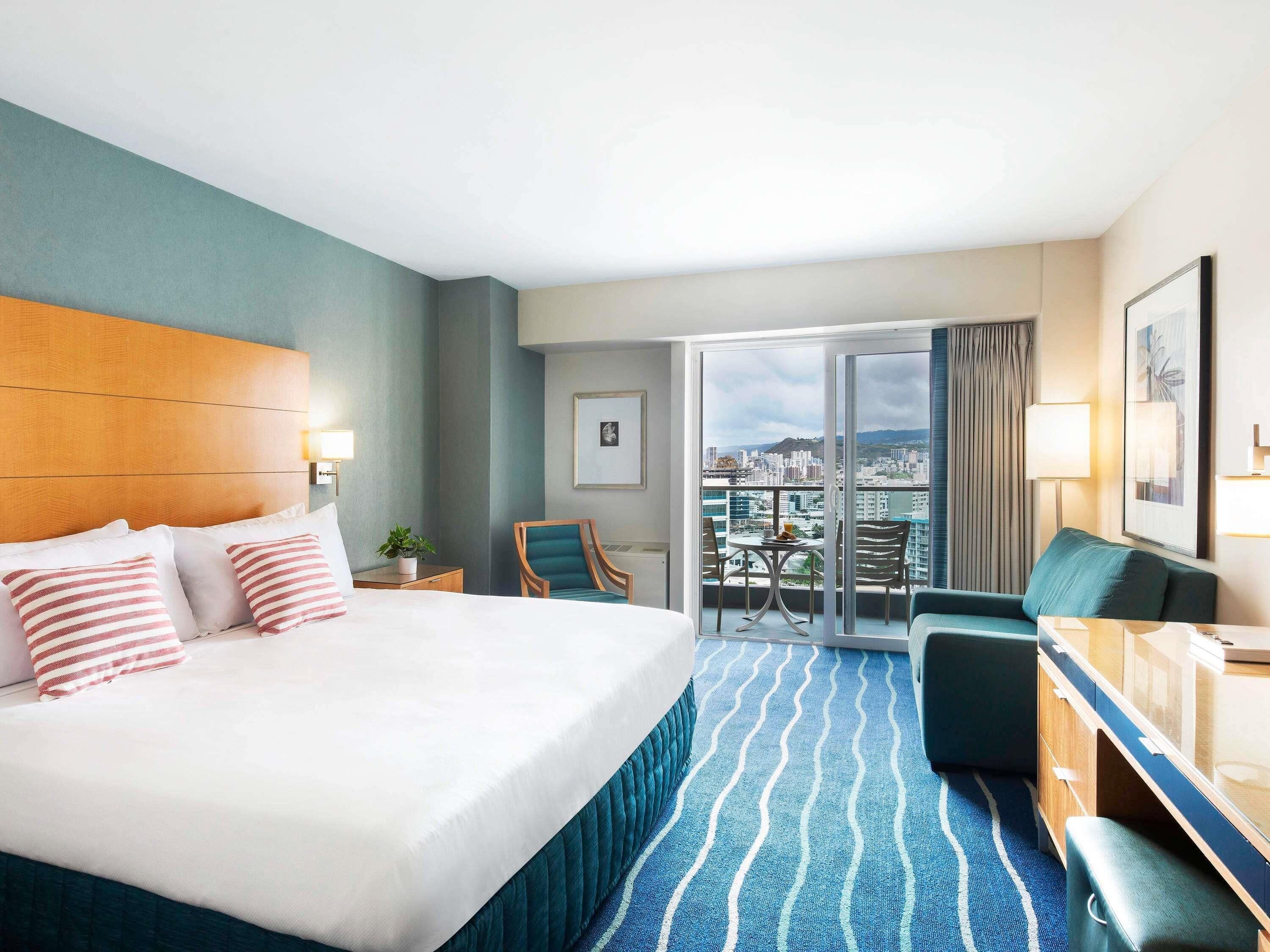 A hotel room with a large bed, a desk, a chair, a balcony with a city view, and a sliding glass door.