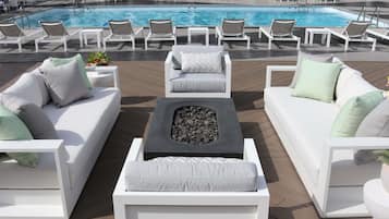 A poolside area with white lounge chairs, a central fire pit, and a pool in the background.