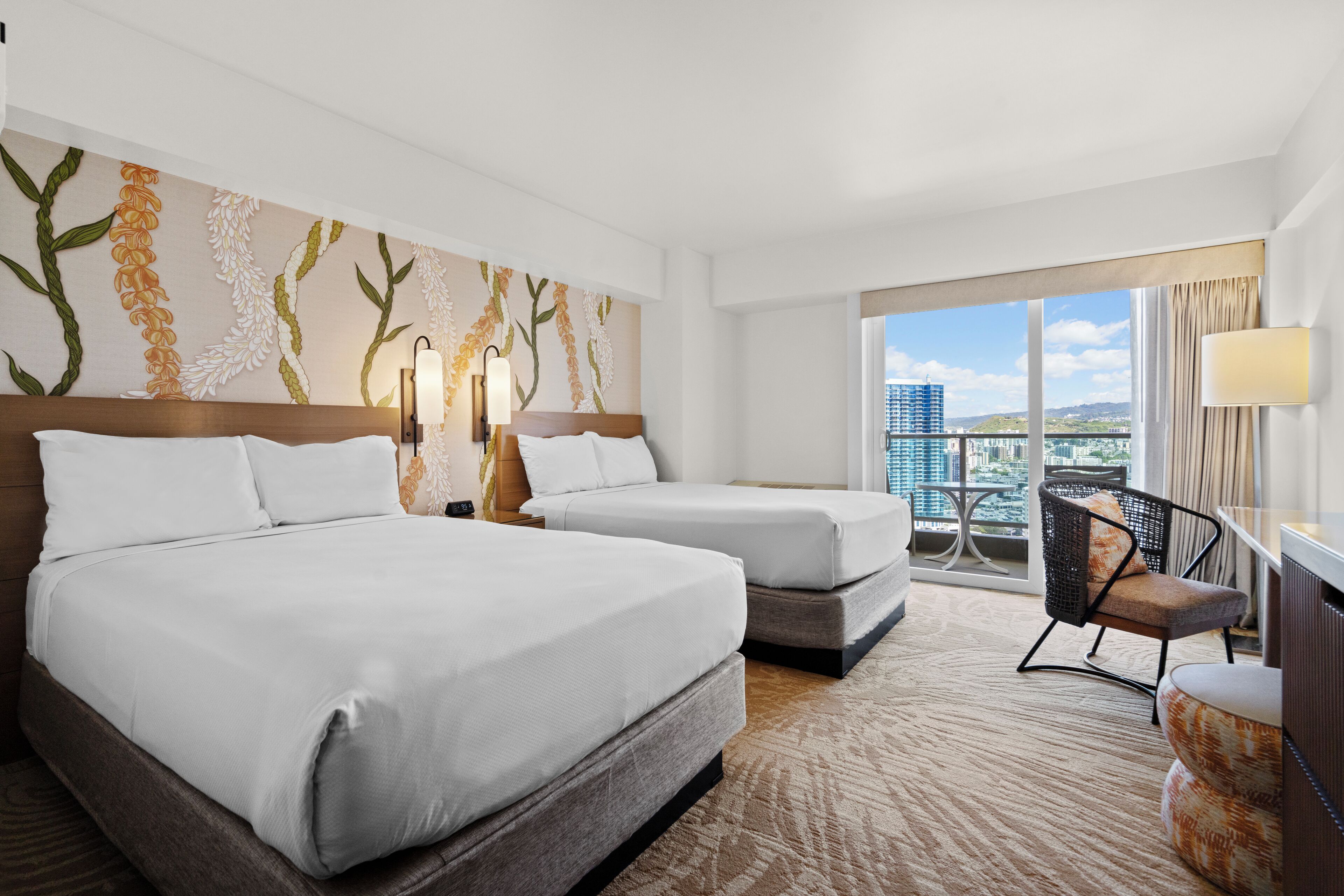Habitación de hotel con dos camas, un ventanal amplio con vistas a la ciudad, un muro decorativo y una zona de estar con una silla y reposapiés.