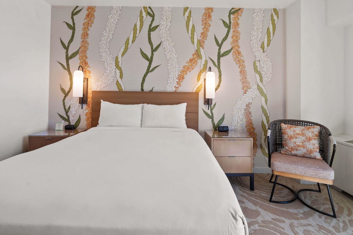 Habitación de hotel con cama, dos mesitas de noche con lámparas, una silla y una pared con estampado floral.
