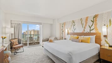 Habitación de hotel con una cama grande, un balcón con vistas a la ciudad, una zona de estar con una silla y una mesa, y una pared decorativa con motivos vegetales.