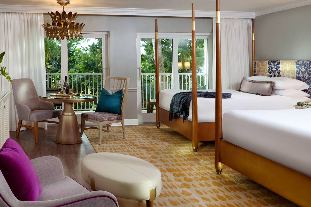 Une chambre d’hôtel comprenant un lit, une chaise, une table avec un vase et des verres à vin, un balcon donnant sur un paysage verdoyant et un luminaire au plafond.