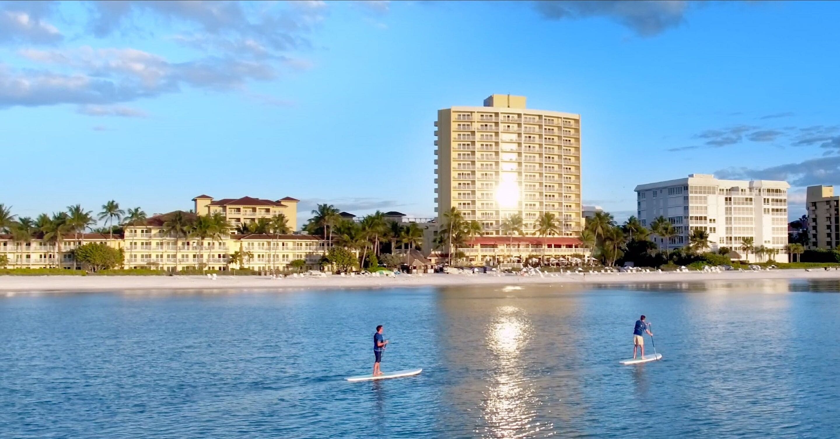 A beachfront hotel with a pool, palm trees, and a beach in the background.