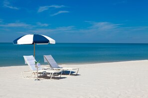 Une plage avec deux chaises longues blanches sous un parasol bleu et blanc, donnant sur l’océan.