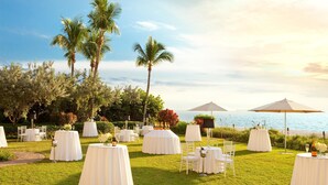 Un salón de bodas al aire libre con mesas redondas, sillas y sombrillas, con palmeras de fondo y vista al mar.