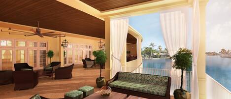A spacious lounge area with a large sofa, multiple chairs, and a ceiling fan. A large window offers a view of a body of water and palm trees.