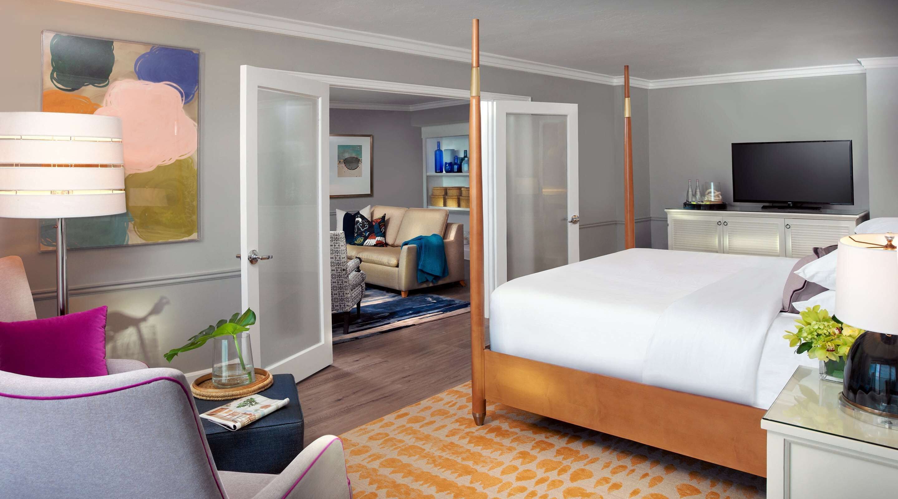 A modern hotel room with a large bed, a television, and a dining area visible through an open door.