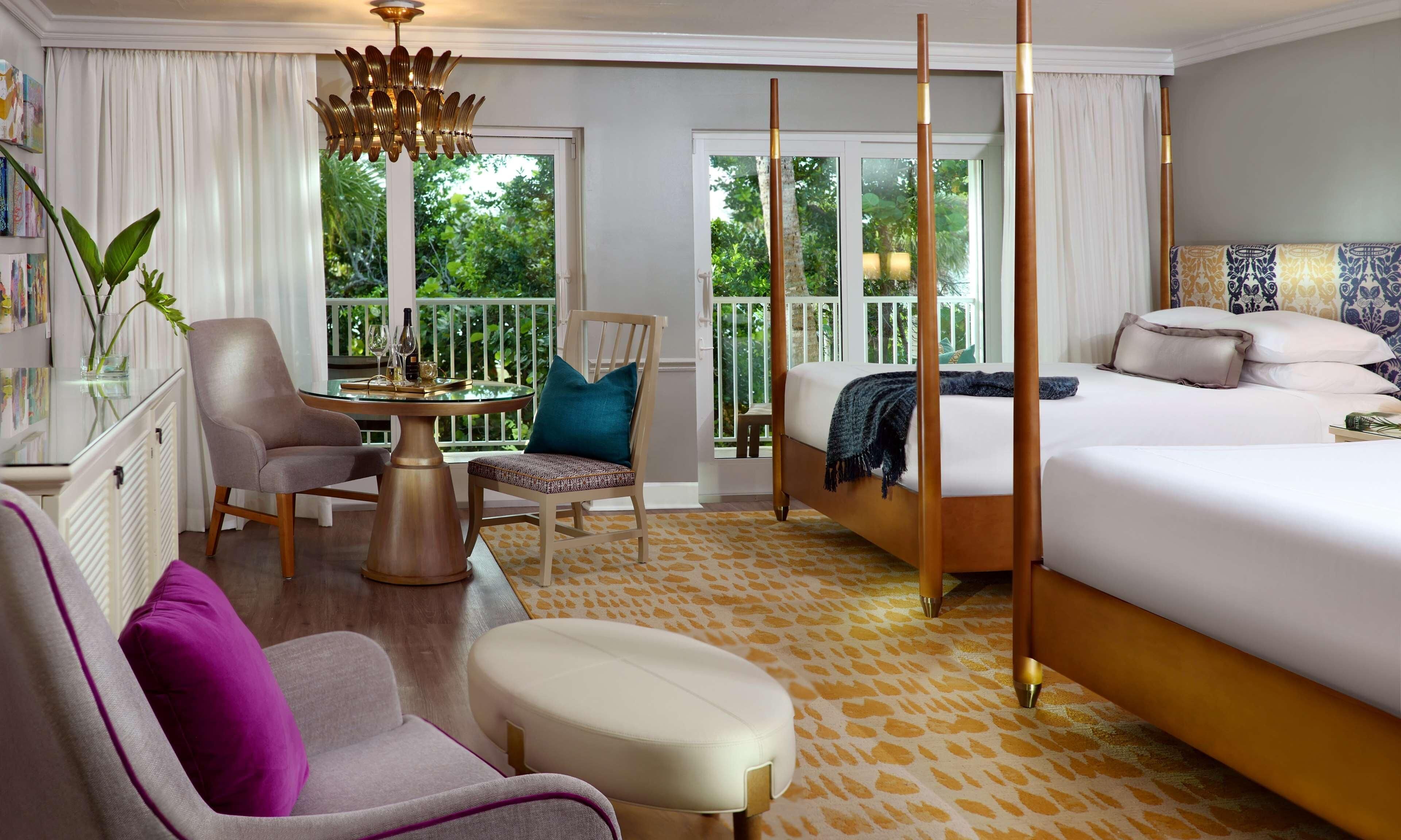 Une chambre d’hôtel avec un lit, une chaise, une table agrémentée d’un vase et de verres à vin, un balcon donnant sur un paysage verdoyant et une moquette à motifs.