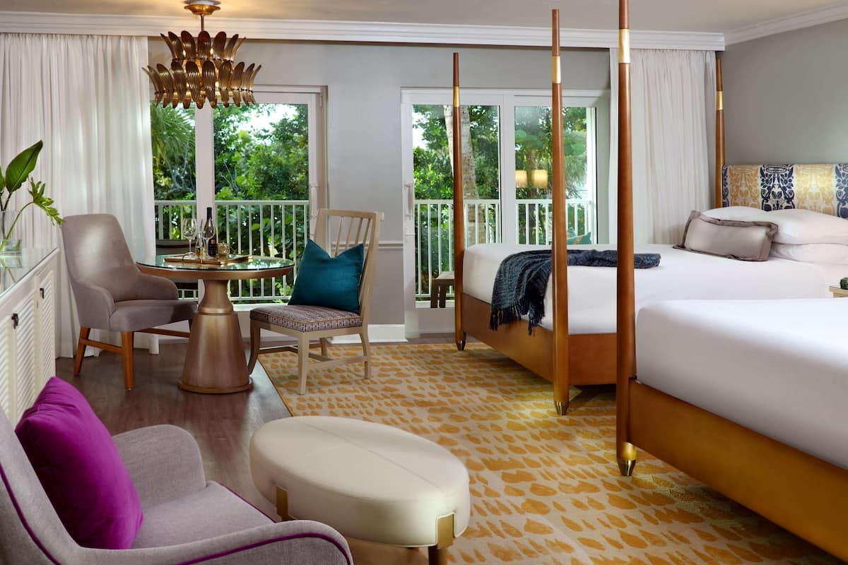 A hotel room with a bed, a chair, a table with a vase and wine glasses, a balcony with a view of greenery, and a patterned carpet.