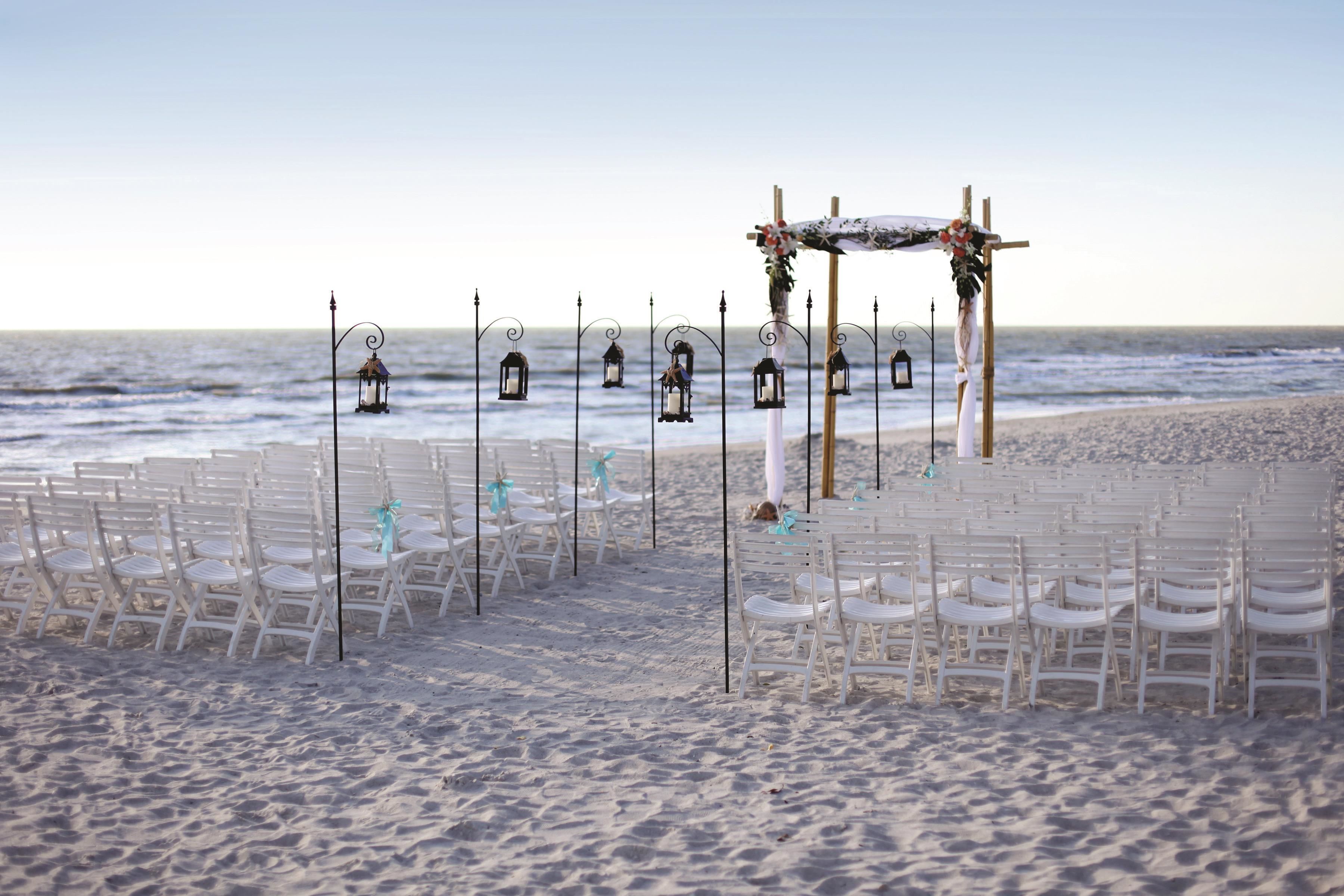 A beach wedding setup with rows of white chairs, a wooden arch, and lanterns.