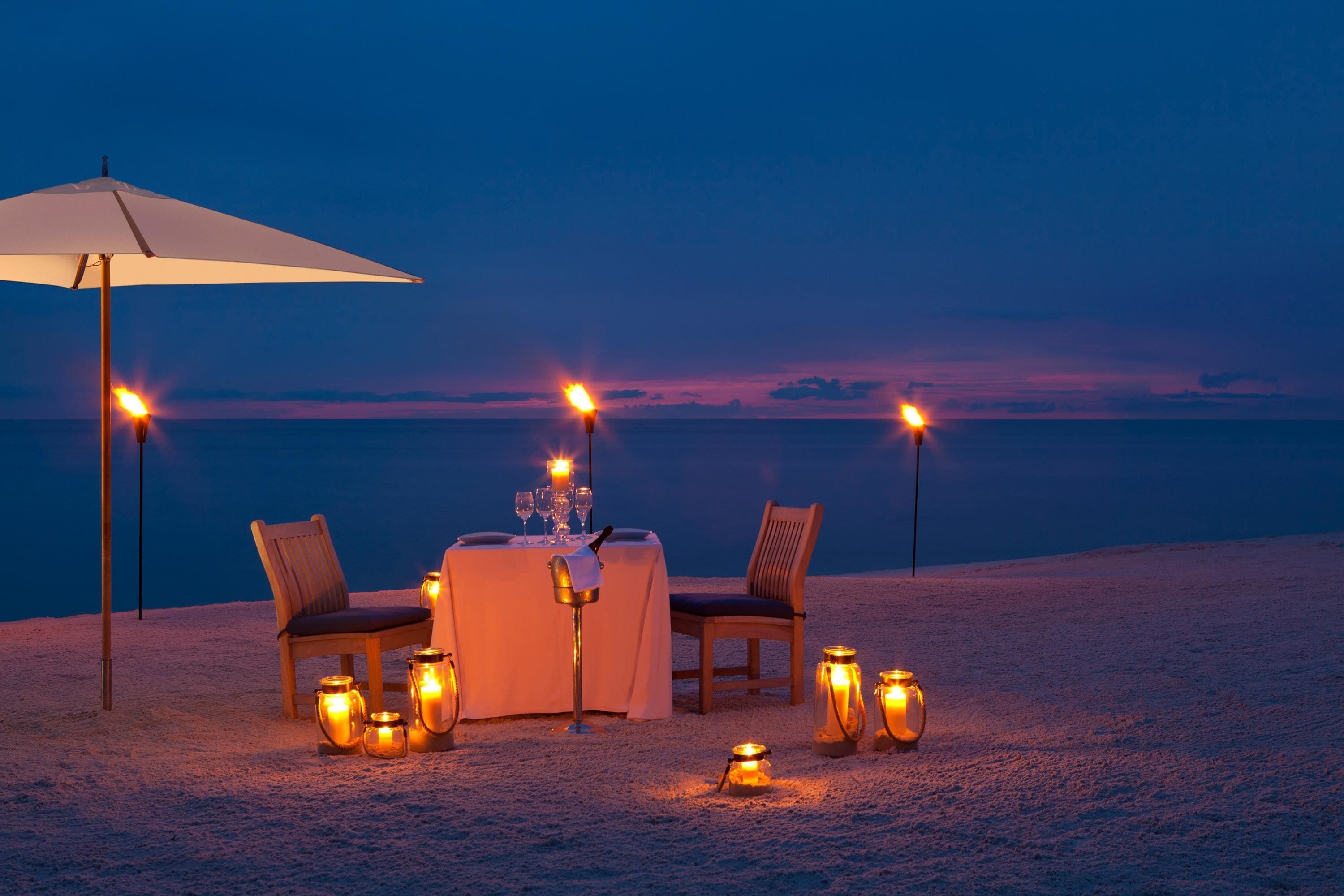Disposição para refeições à beira-mar, com mesa, cadeiras, guarda-sol e velas.