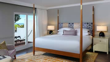A four-poster bed with a white bedspread, two bedside tables with lamps, a chair with a cushion, and a view of the outdoors through a sliding door.
