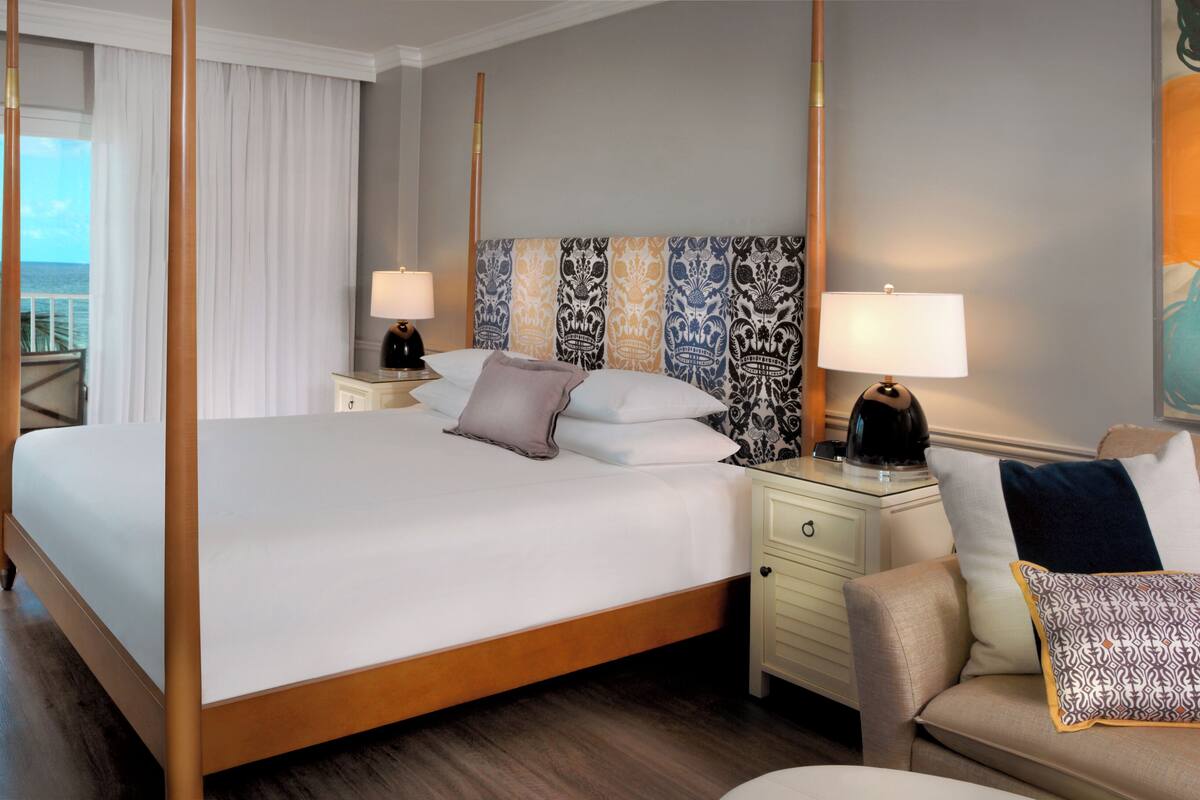 Une chambre spacieuse avec un grand lit, deux tables de chevet, une commode, une chaise et une vue sur l’océan grâce à de grandes fenêtres.