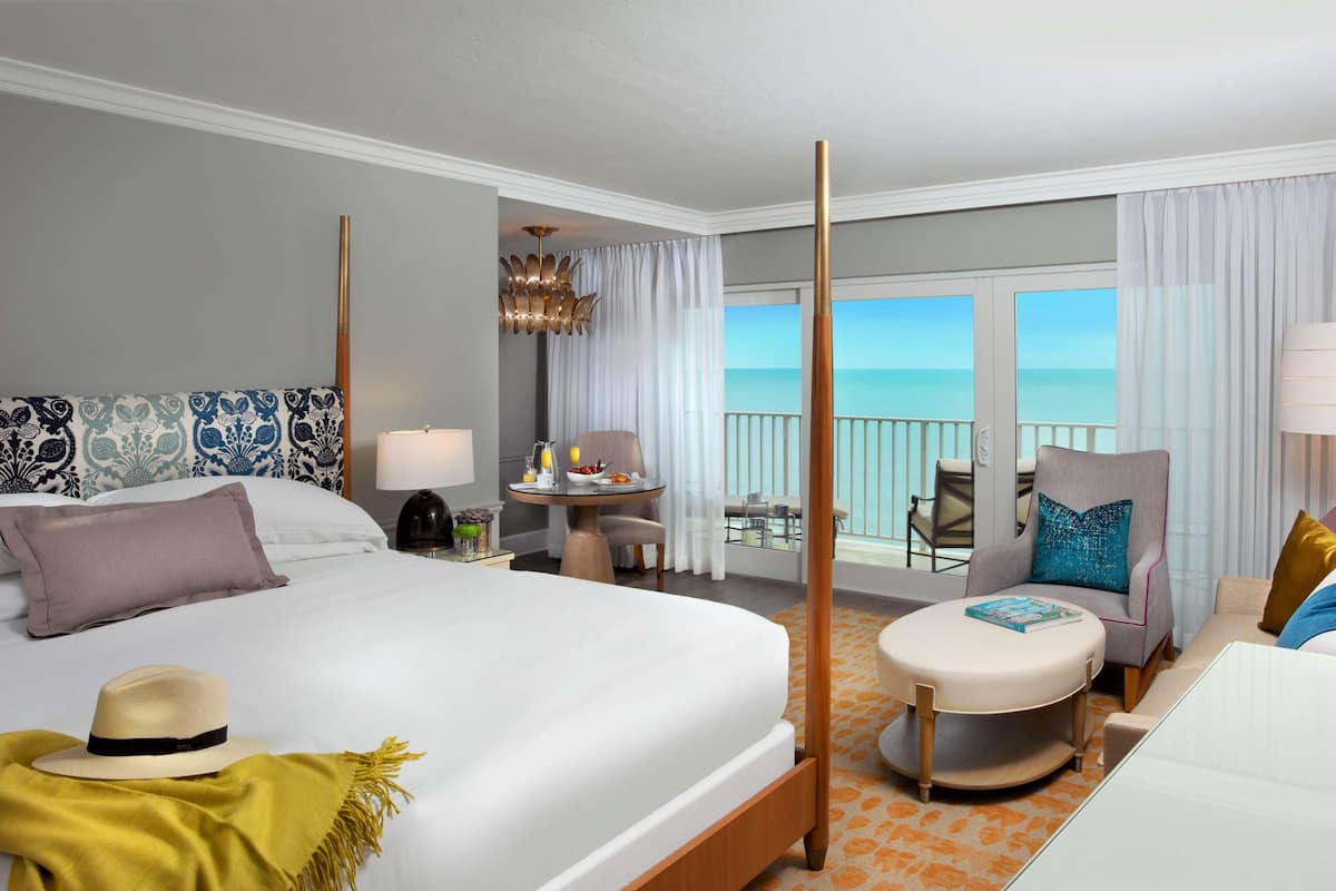 A hotel room with a bed, a balcony with a view, a small table with a lamp, and a chair.