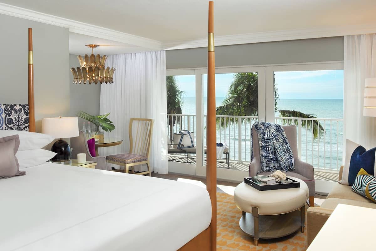 A hotel room with a large bed, a chair, a small table, a lamp, and a view of the ocean.
