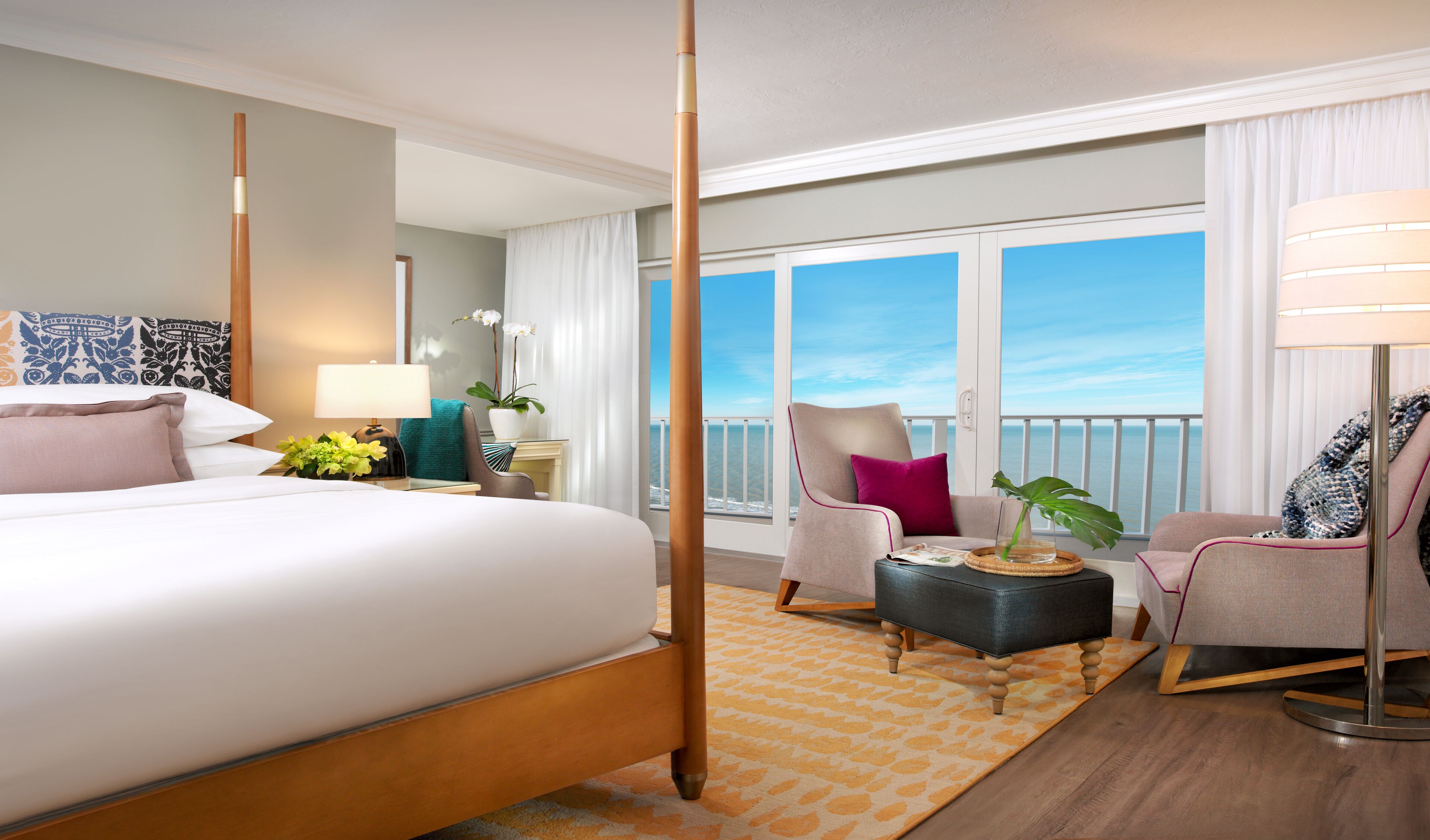 A hotel room with a four-poster bed, a sitting area with a chair and ottoman, a small table with a plant, and a view of the ocean through large windows.