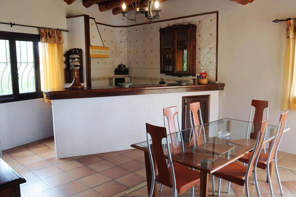 Un comedor con mesa de vidrio y sillas metálicas, una cocina con mostrador de madera y un ventanal con cortinas.