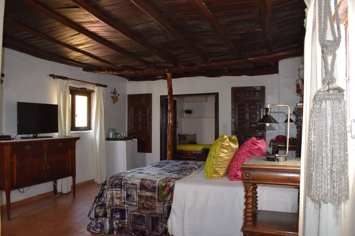 Una habitación con techo de madera, una cama con colcha, un escritorio con lámpara y un televisor sobre un mueble.