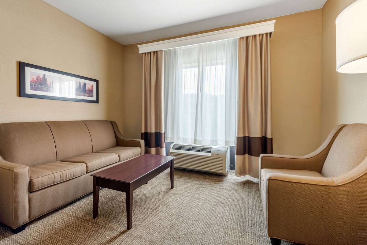 Ein Hotelzimmer mit einem beigen Sofa, einem Holztisch, einem beigen Sessel und einem großen Fenster mit transparenten Vorhängen.