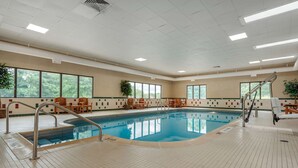 A large indoor swimming pool with a tiled floor, wooden chairs, and a staircase leading to a balcony.