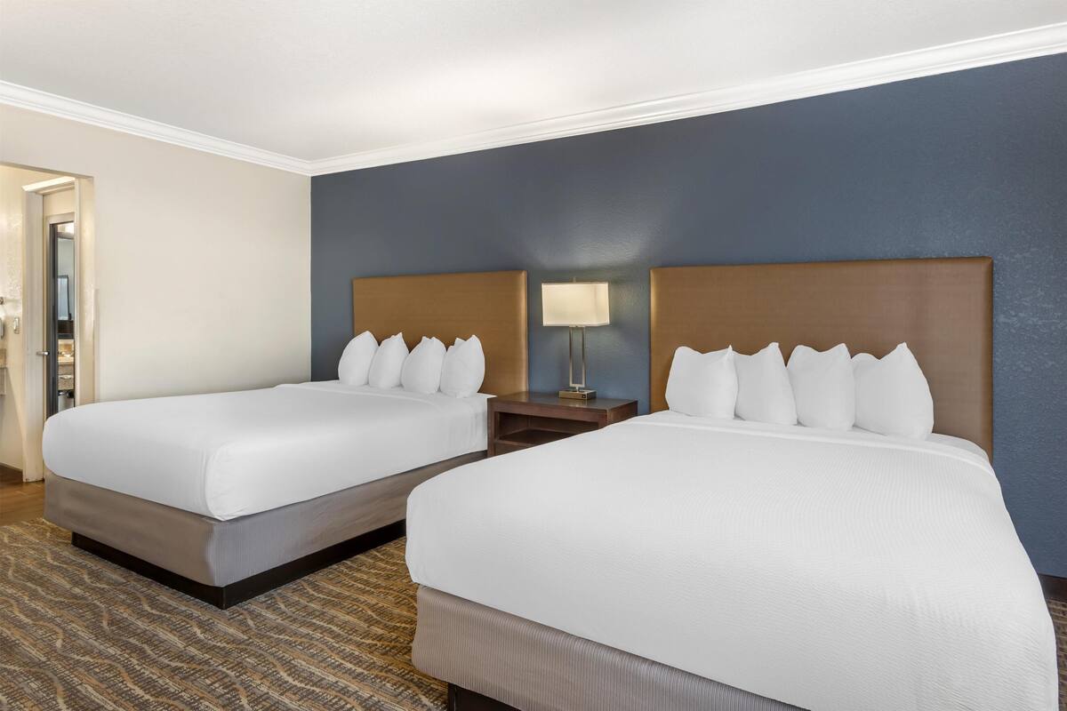 A hotel room with two beds, a nightstand with a lamp, and a view of the hallway.