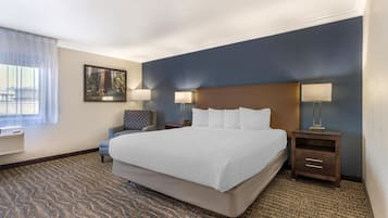 Quarto de hotel com uma cama grande, duas mesas de cabeceira com abajures, uma cadeira, um quadro na parede e um tapete estampado.