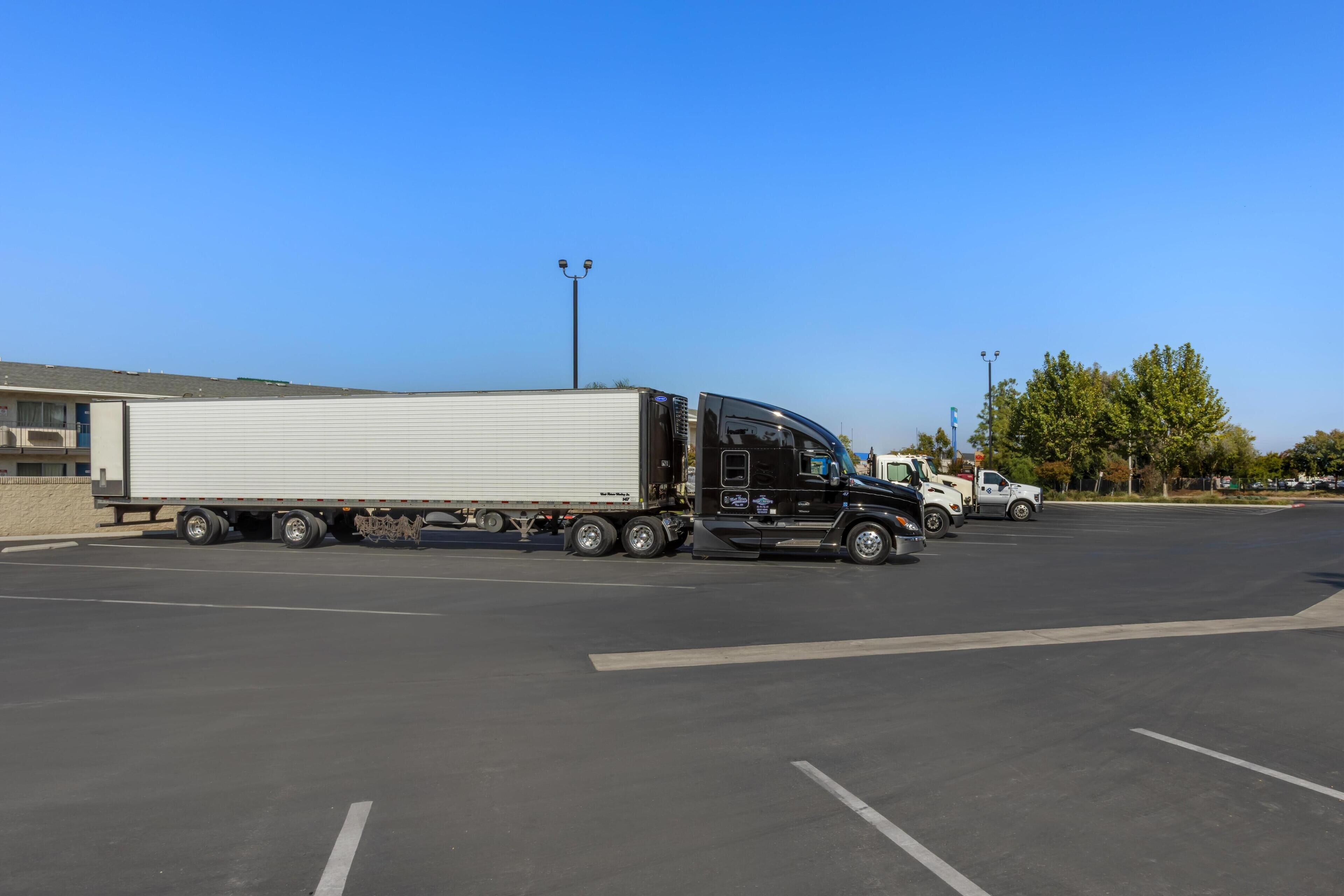 A parking lot with a black semi-truck and a white trailer, a white utility truck, and a building with a flat roof in the background.