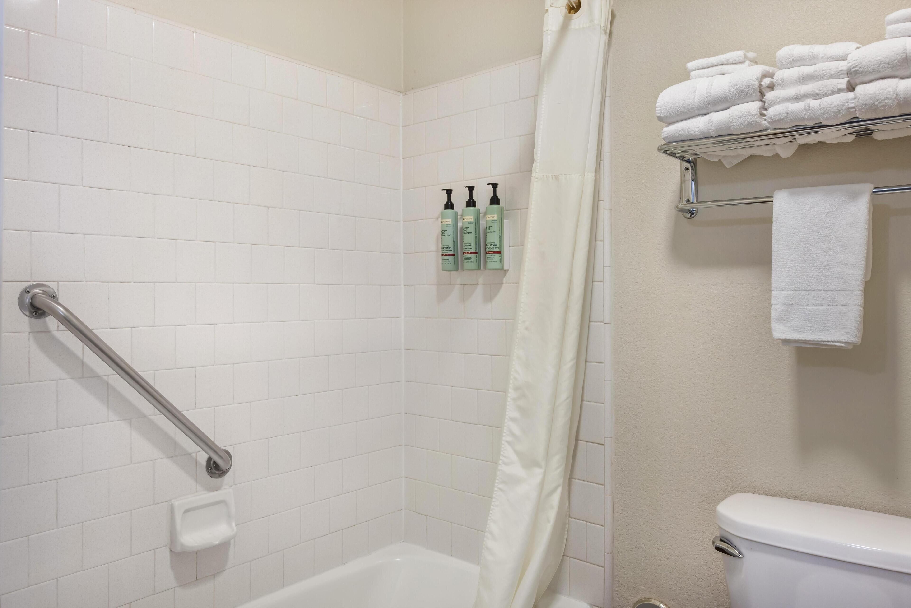 A bathroom with white tiled walls, a white bathtub, a shower curtain, a grab bar, a shelf with toiletries, and a towel rack with folded towels.
