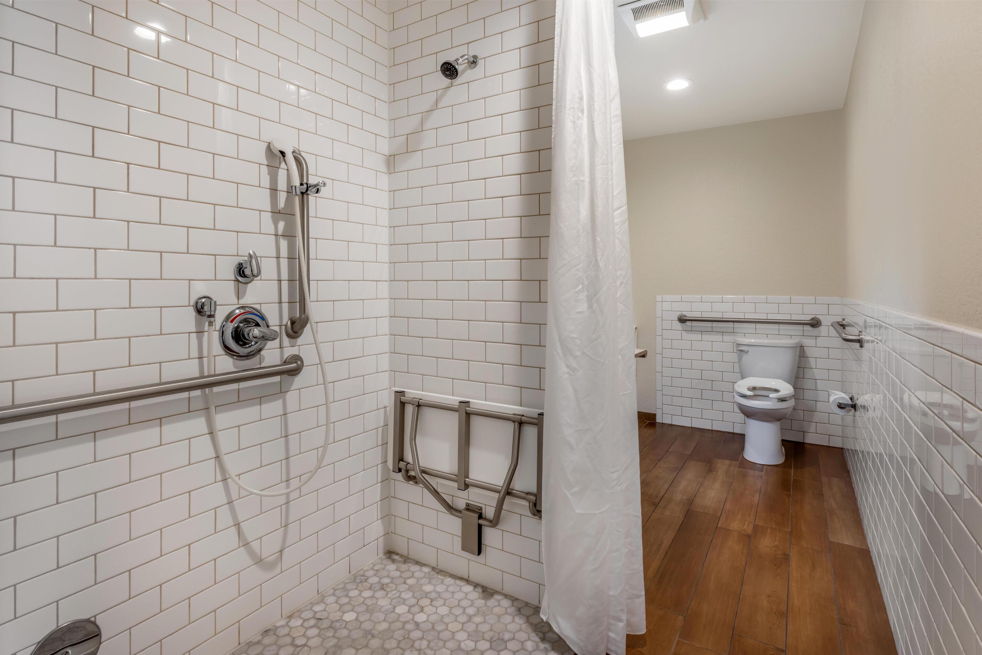 A bathroom with white tiled walls, a shower with a grab bar, a toilet, and a wooden floor.