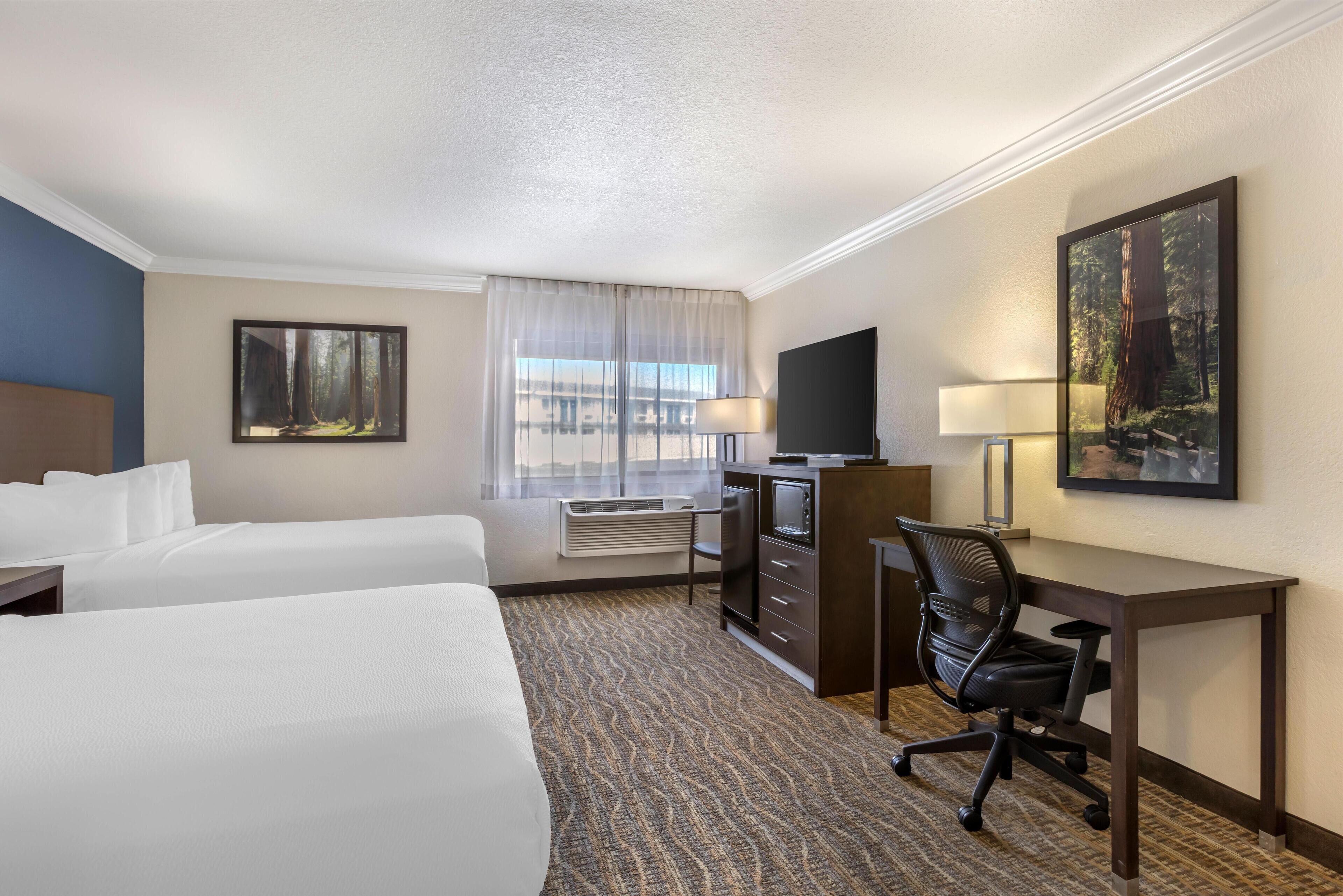 A hotel room with two beds, a desk with a chair, a TV, and a large window with curtains.