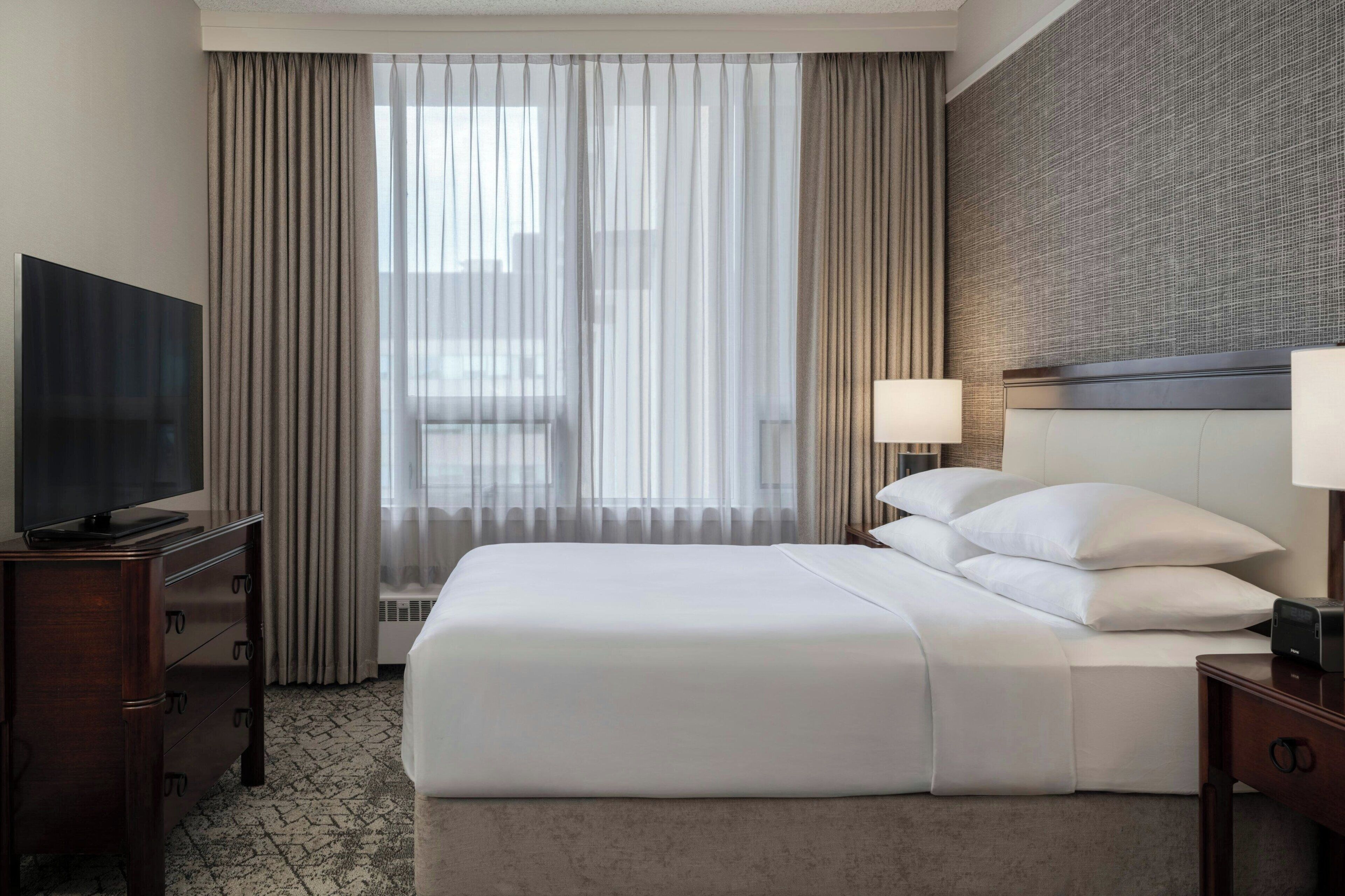 A hotel room with a large bed, a television on a wooden dresser, and a window with sheer curtains.