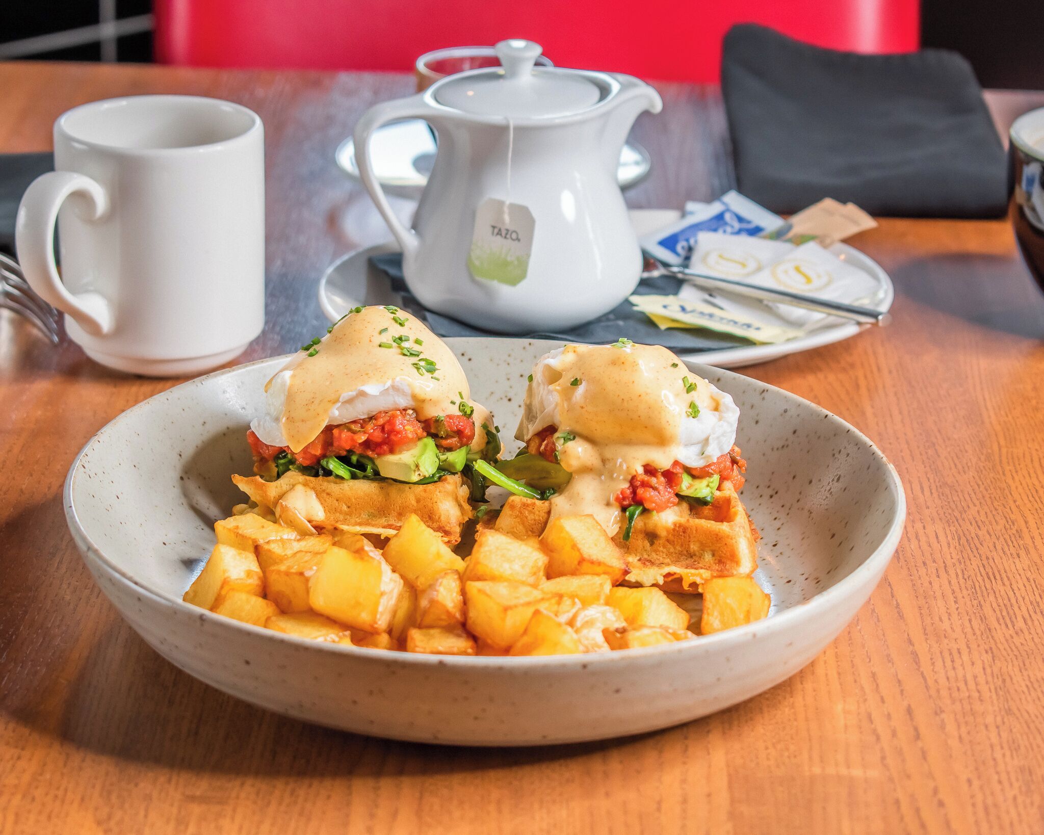 Un plato de desayuno con huevos Benedictinos, croquetas de patata y una tetera con la etiqueta "TAZO" sobre una mesa.