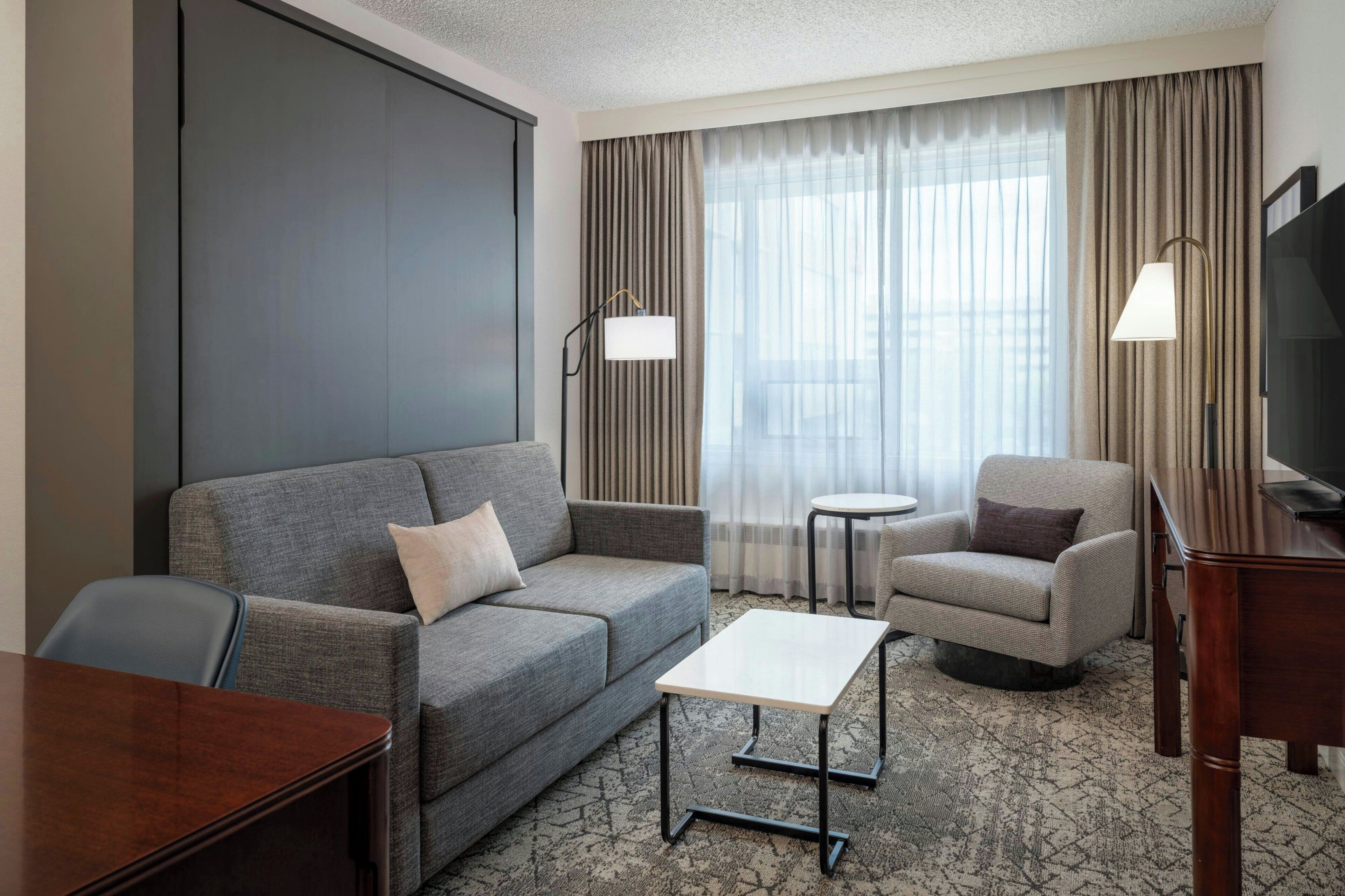 A hotel room with a grey sofa, a small round table, a TV stand, and a large window with curtains.