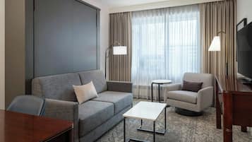 A hotel room with a grey sofa, a small round table, a TV stand, and a large window with curtains.