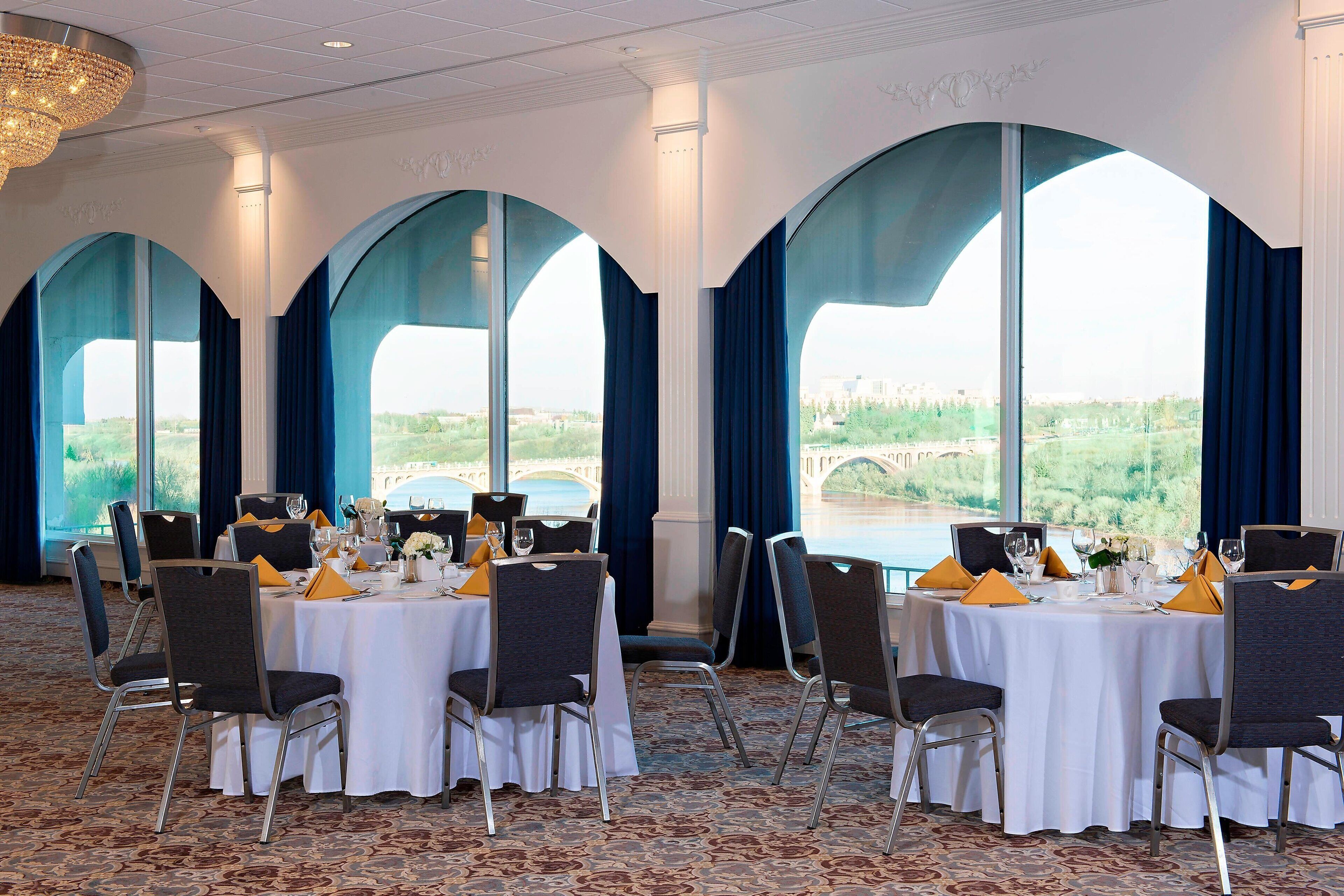 A banquet hall with round tables set for dining, large arched windows, and a chandelier.