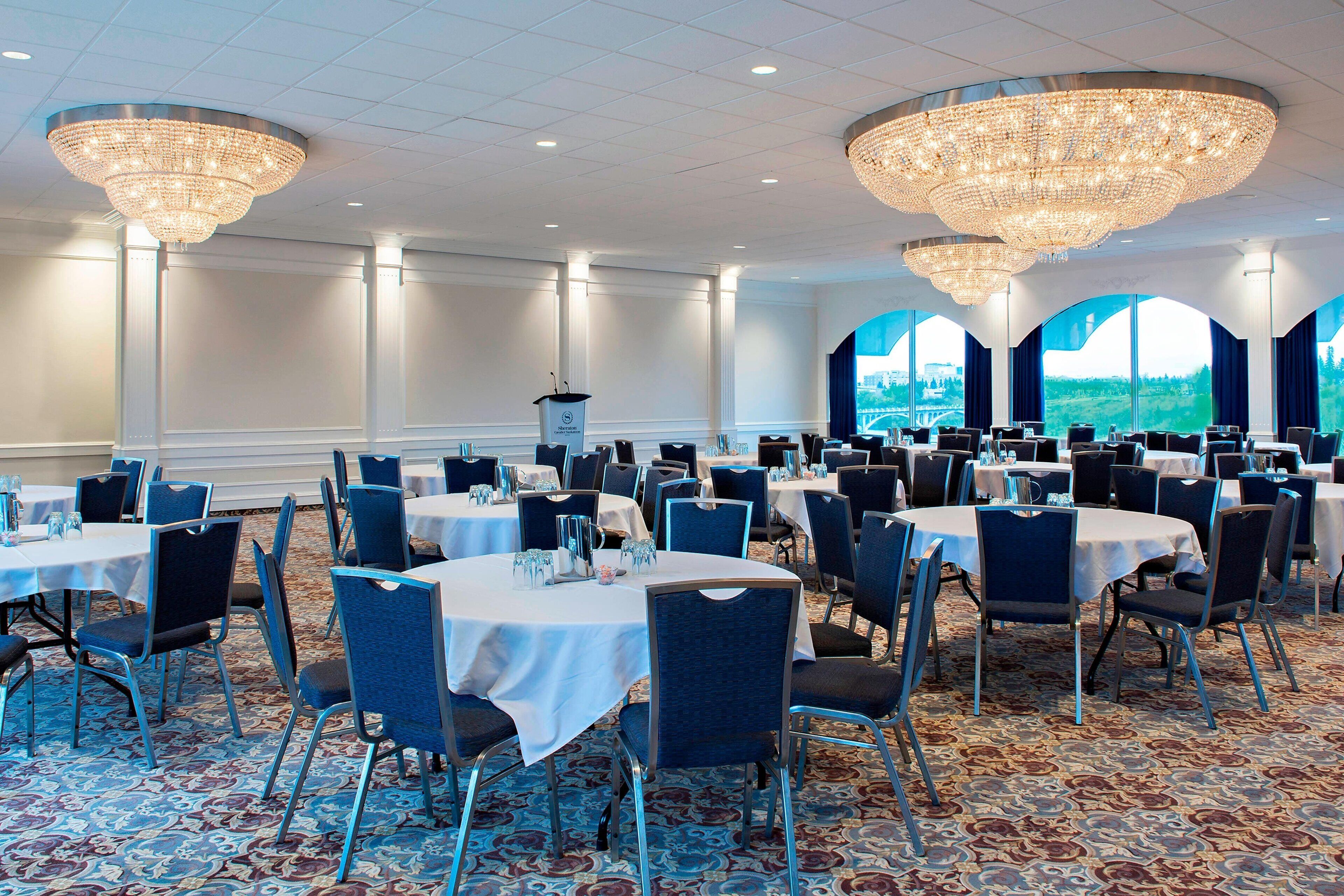 A banquet hall with round tables, chairs, chandeliers, and large windows offering a view of the outdoors.