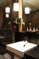 Un baño moderno con lavamanos blanco, baldosas oscuras y dos lámparas colgantes.