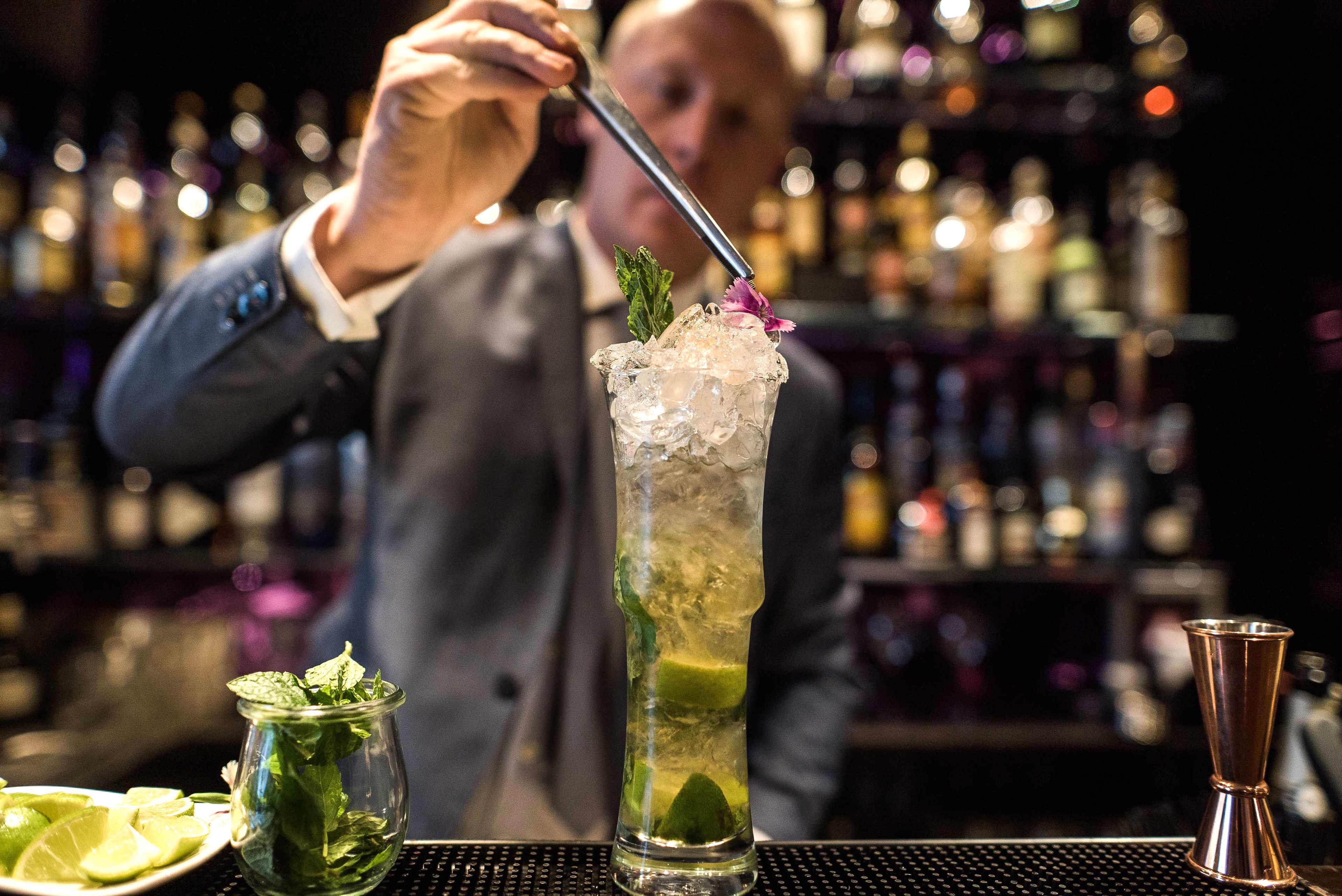 A bartender preparing a cocktail with a tall glass filled with ice, garnished with mint and a flower.
