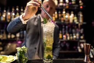 Un barman preparando un cóctel en un vaso alto lleno de hielo, decorado con menta y una flor.