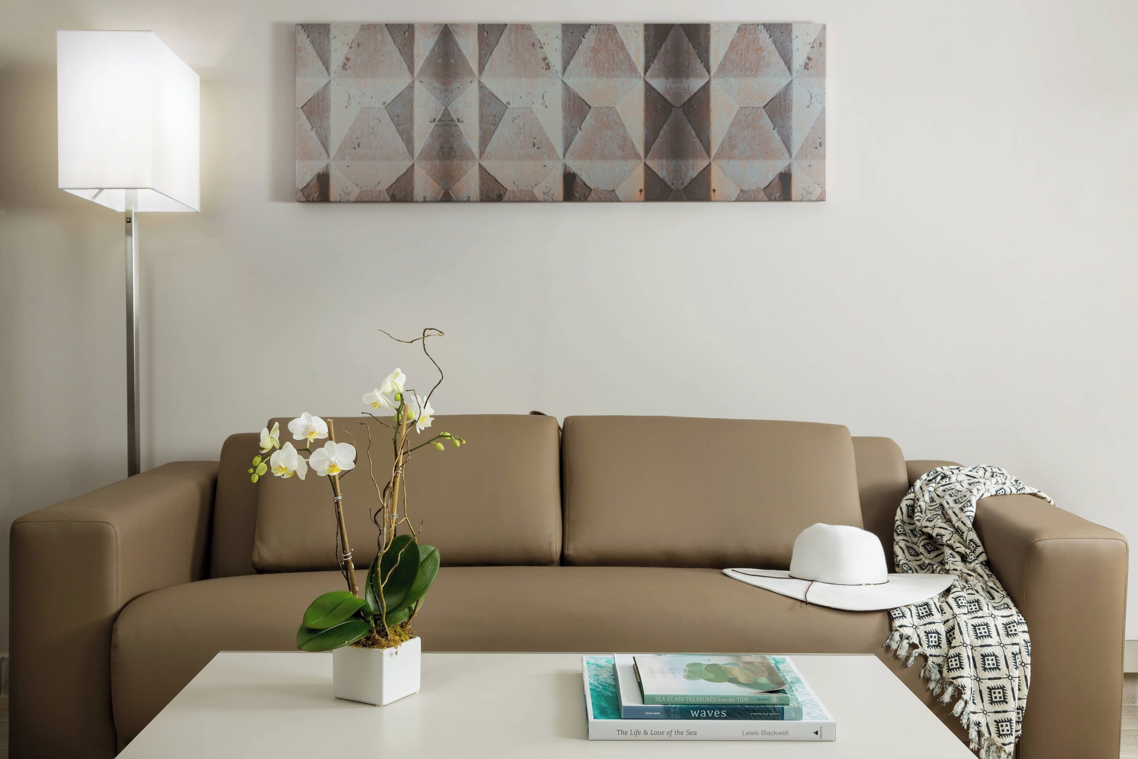 A modern living room with a beige sofa, a white coffee table, a potted orchid, a white hat, and a book titled "The Life & Love of the Sea" by Lewis Blackwell.
