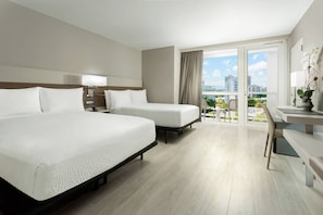 Ein Hotelzimmer mit zwei Betten, einem Schreibtisch und einem Balkon mit Blick auf Gebäude und Grünflächen.
