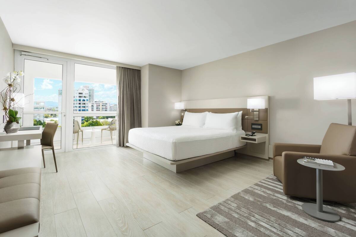 Ein modernes Hotelzimmer mit einem großen Bett, einem Schreibtisch, einem Sessel, einer Couch und einem Balkon mit Stadtblick.