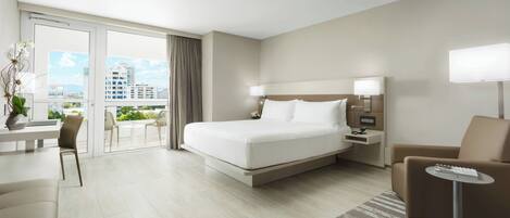 A modern hotel room with a large bed, a desk, a chair, a sofa, and a balcony with a view of the city.