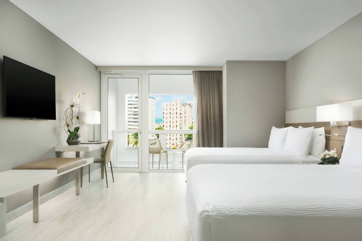 Ein Hotelzimmer mit Bett, Schreibtisch, Stuhl, Fernseher und einem Balkon mit Blick auf Gebäude.