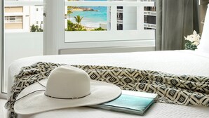 Habitación de hotel con cama, ventana con vista a la playa y edificios, un sombrero y un libro.